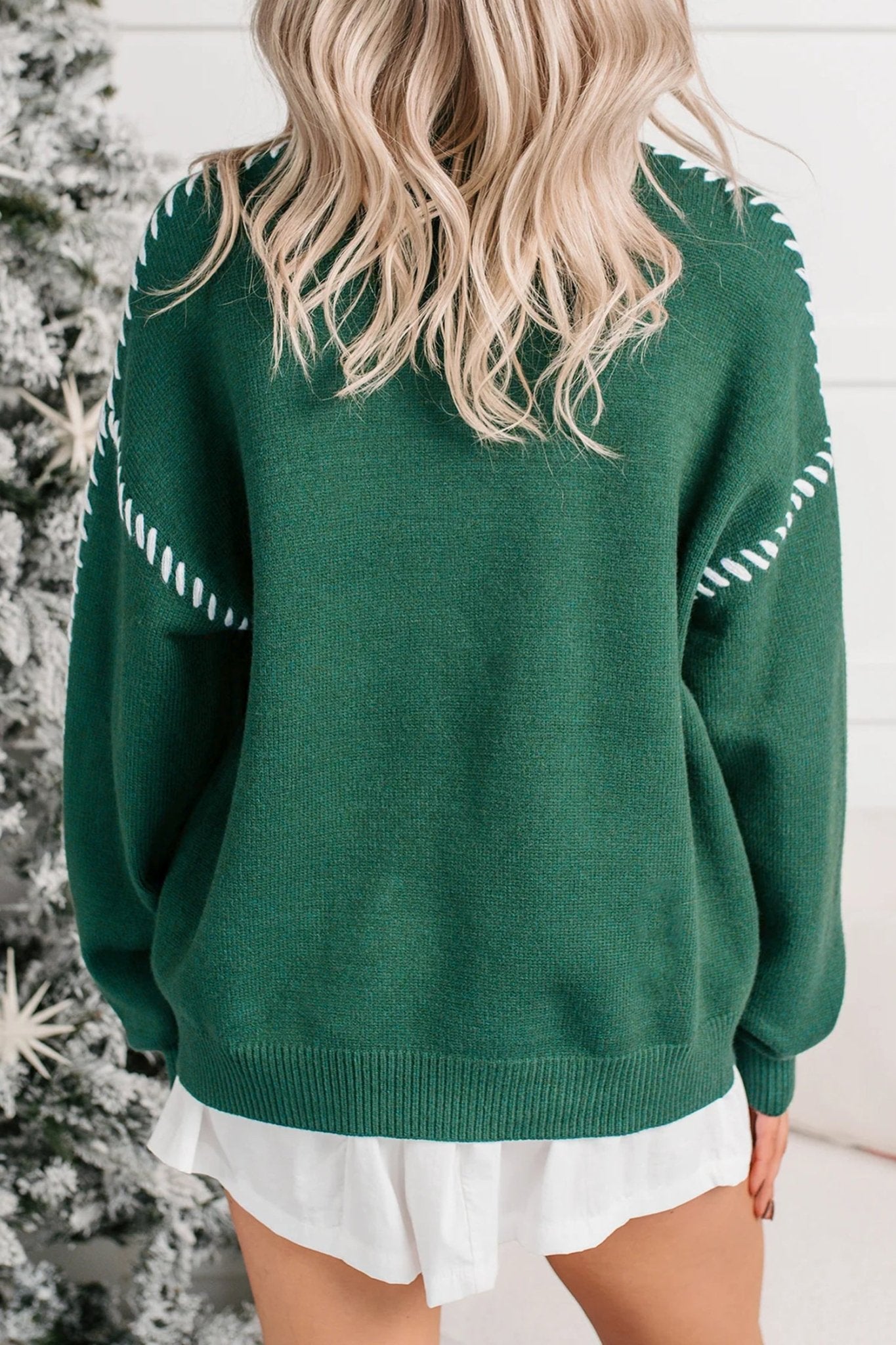 Woman wearing a dark green knit sweater with white stitching and a white skirt.