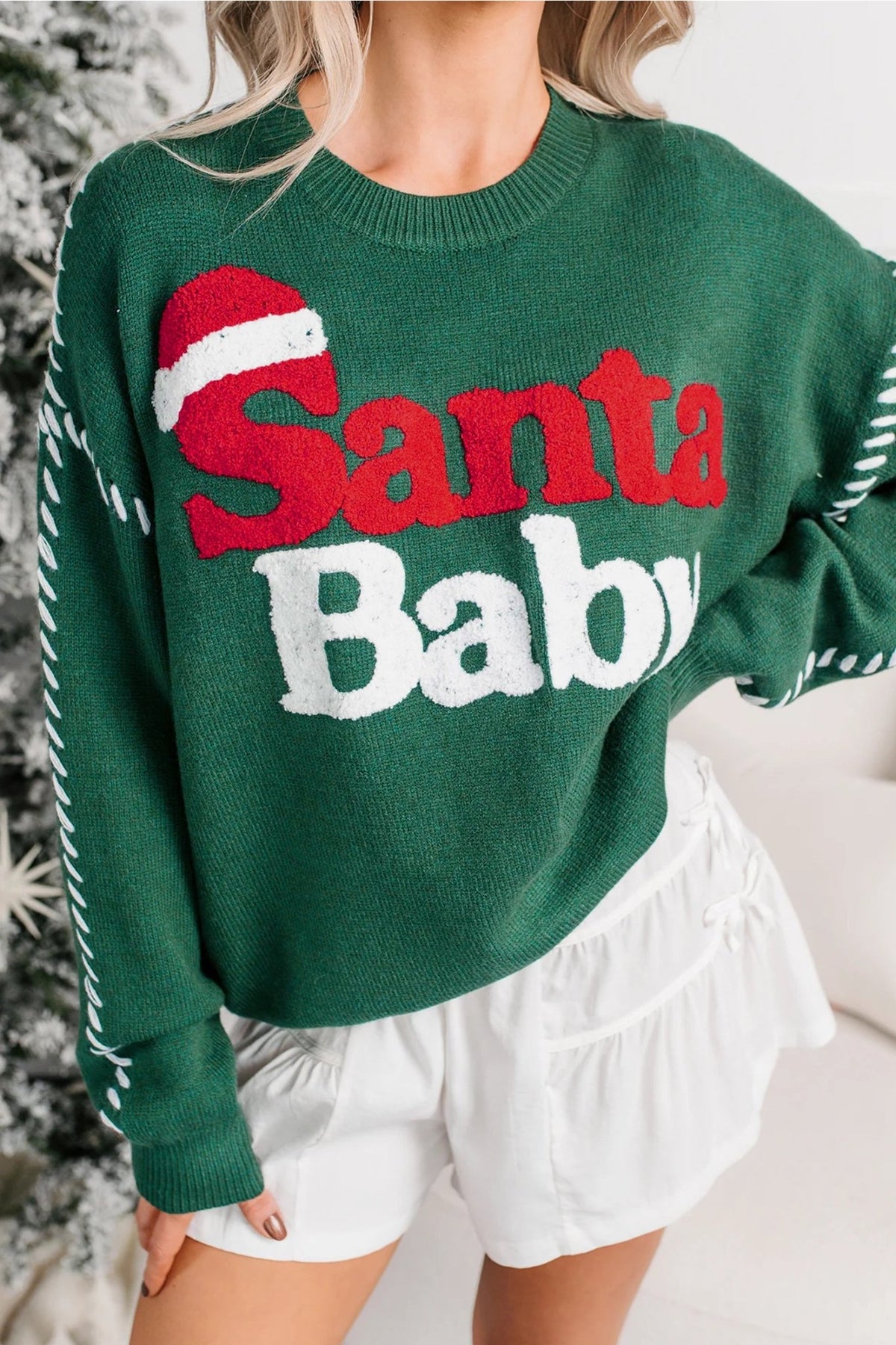 Woman wearing a festive green “Santa Baby” sweater with white shorts.