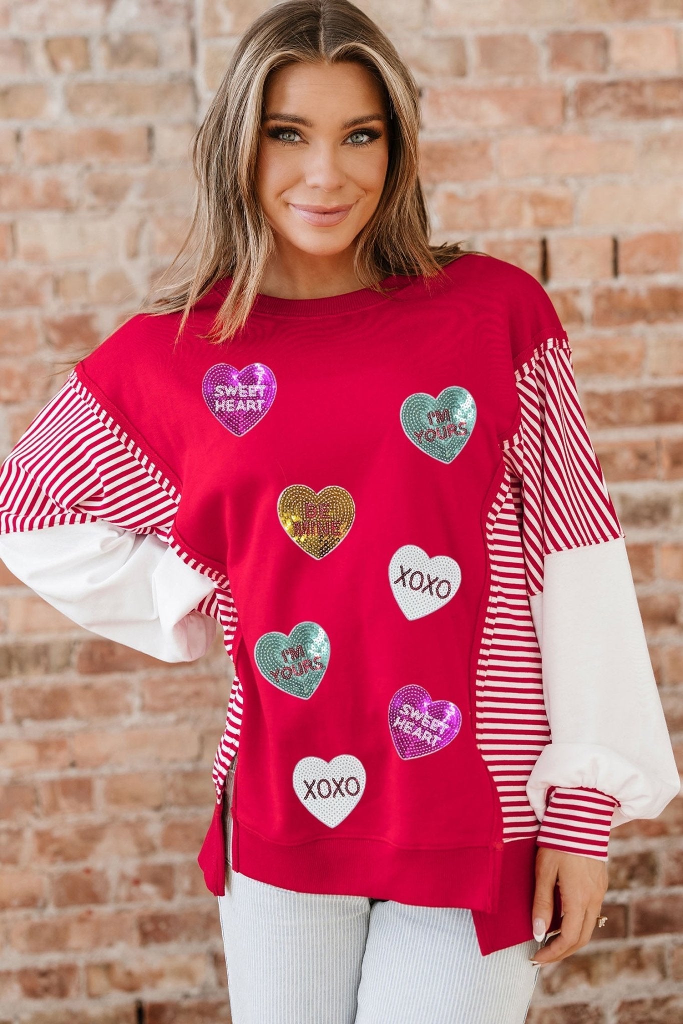 Woman wearing a red sweater with heart-shaped patches against a brick wall.