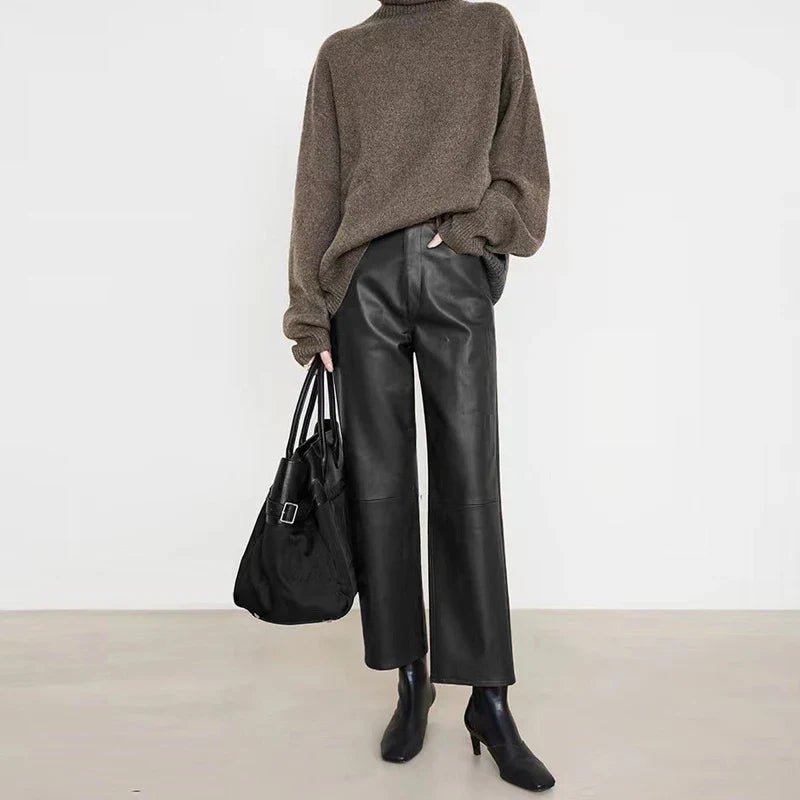 Person wearing a brown sweater and black leather pants holding a black bag against a white background