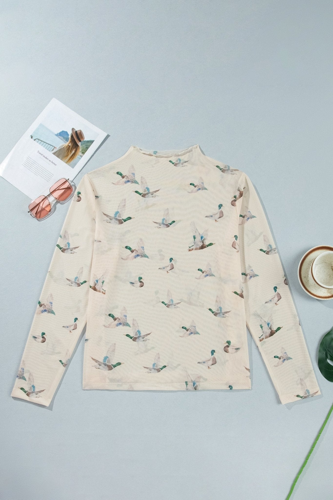 Cream mock-neck top with flying duck print, styled with glasses and magazine.