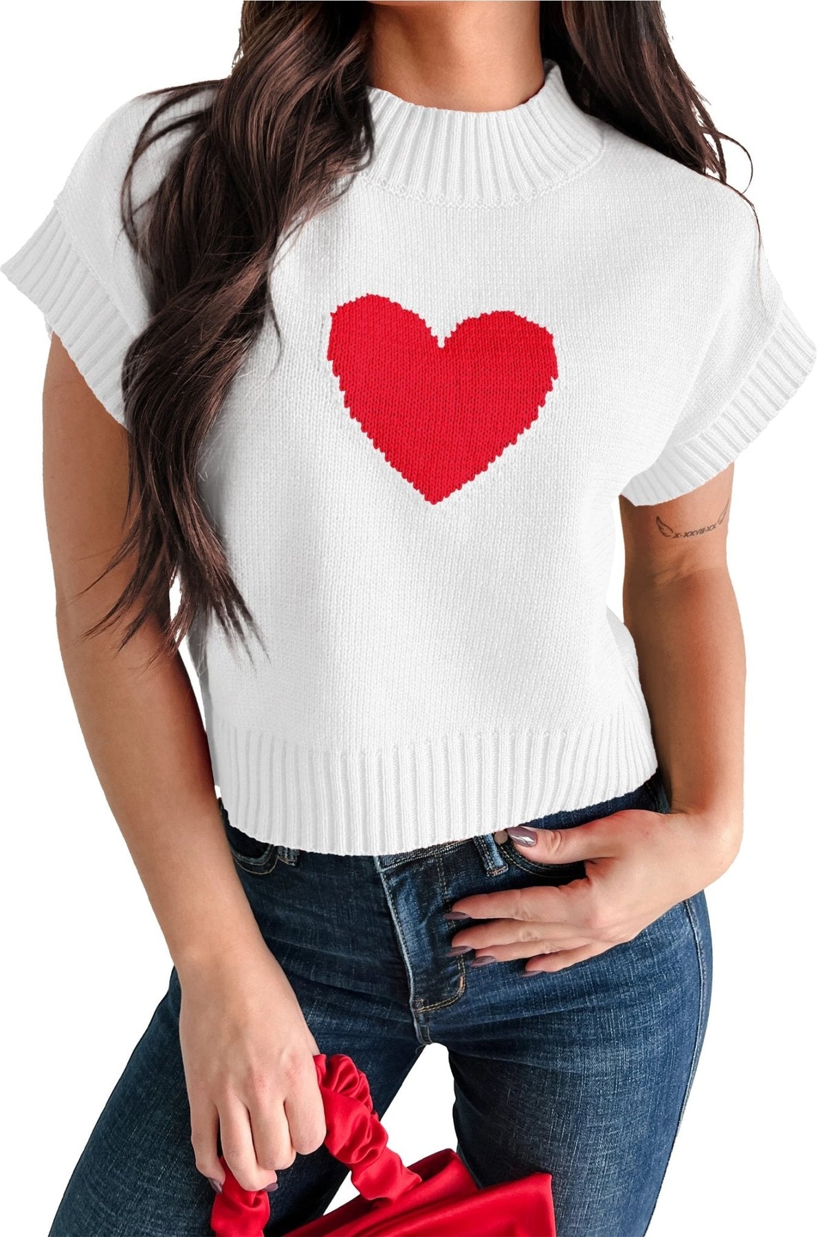White sweater with a red heart design worn by a person, holding a red bag.
