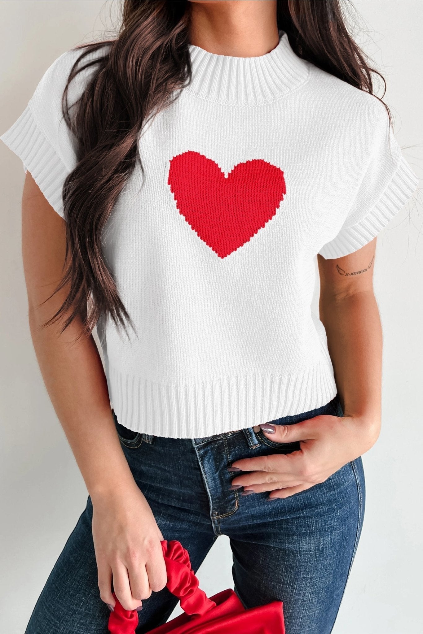 White sweater with a red heart design worn by a person, holding a red bag.