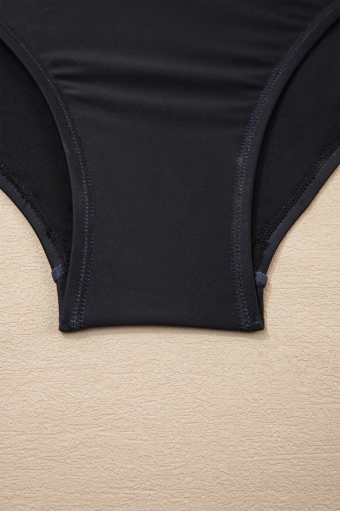 Close-up of a black bikini bottom or underwear leg opening laid flat on a light beige textured surface.