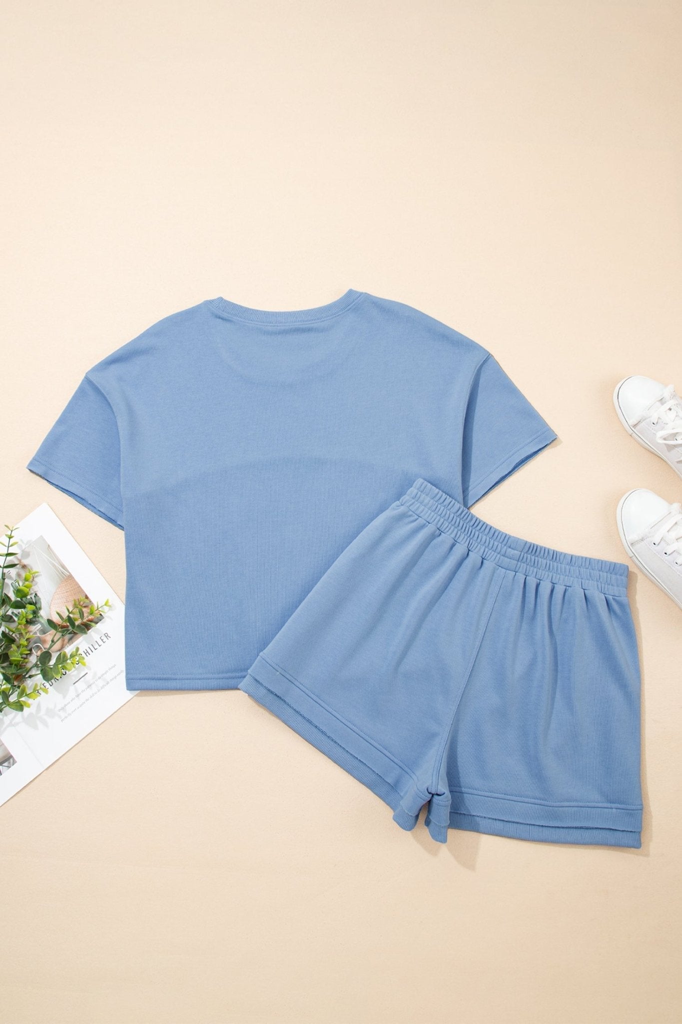 Blue t-shirt and shorts set on a beige surface with a magazine and sneakers in the background.