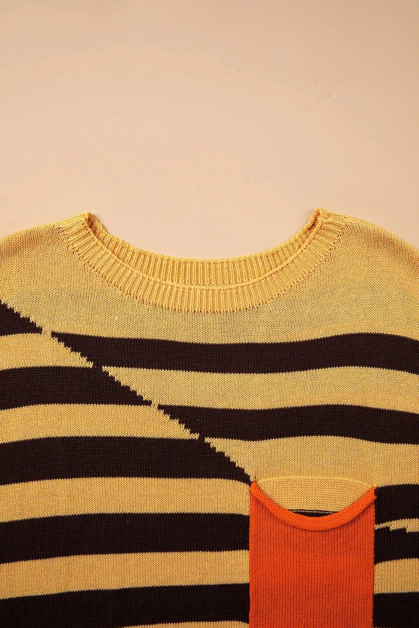 Close-up of black and beige striped knit sweater with contrasting orange pocket on beige background.
