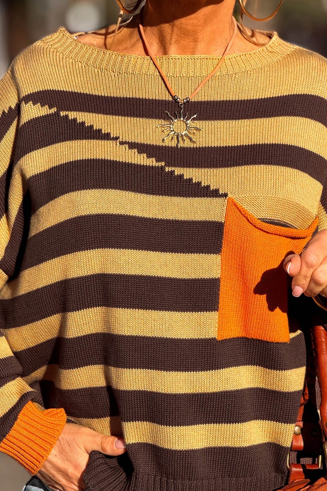 Close-up of a striped knit sweater with orange pocket and cuffs, warm sunlight, jewelry visible.
