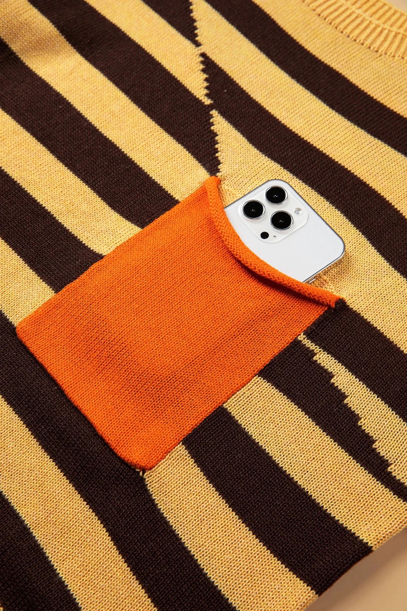 Close-up of mustard and brown striped knit sweater with orange pocket and white smartphone.

