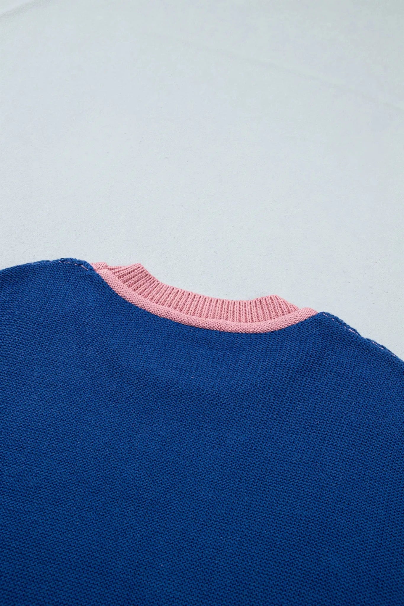 Close-up of a blue knit sweater with a pink ribbed crew neck trim, shown against a plain light background.