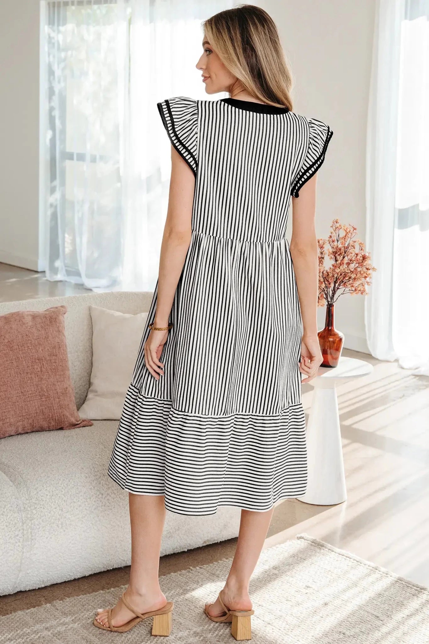 A woman wearing a black and white vertically striped midi dress with ruffled cap sleeves and black trim at the neckline, standing in a bright living room.