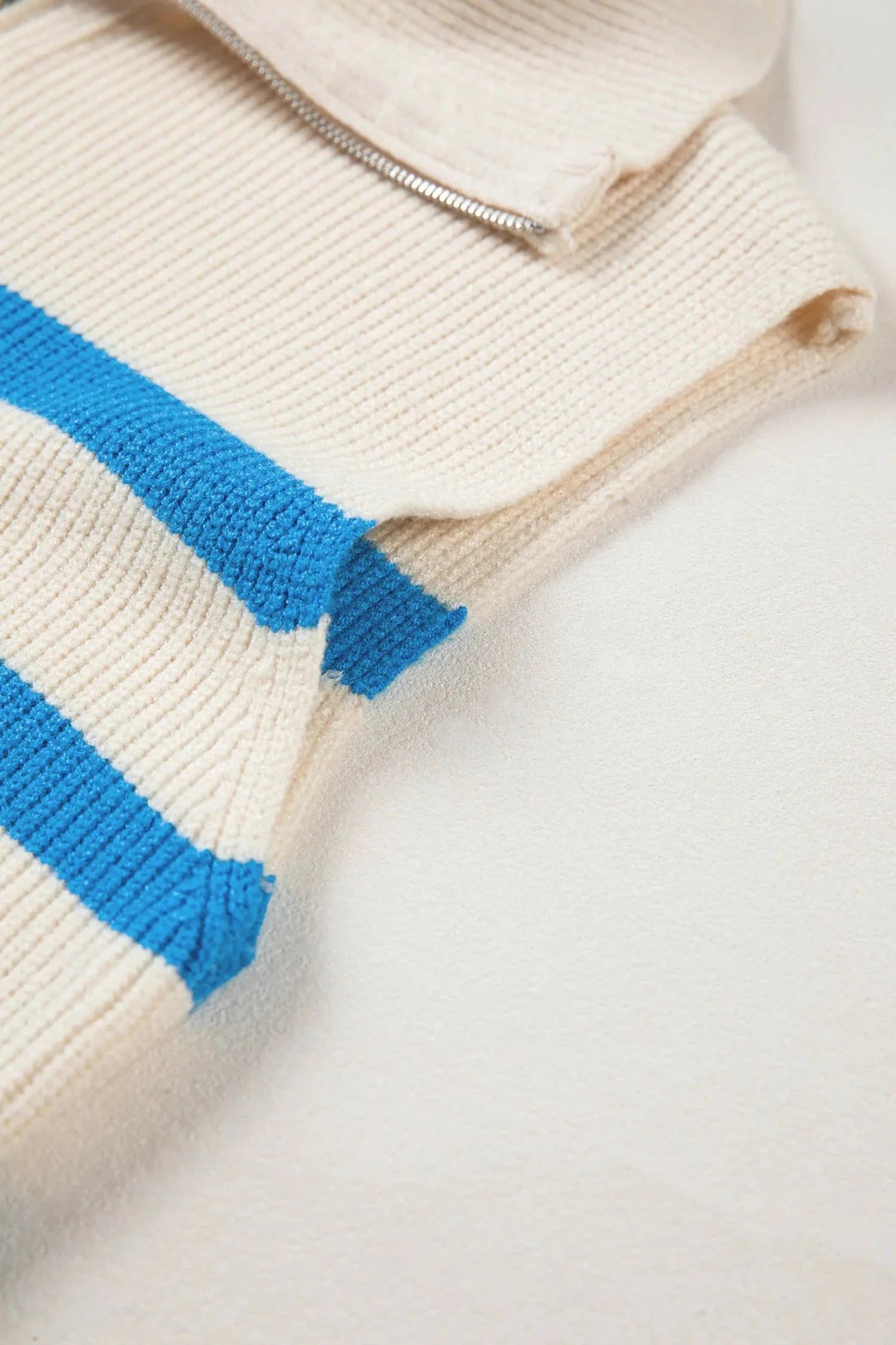 Close-up of a cream-colored ribbed knit garment with bright blue horizontal stripes and a silver zipper, laid flat on a white surface.