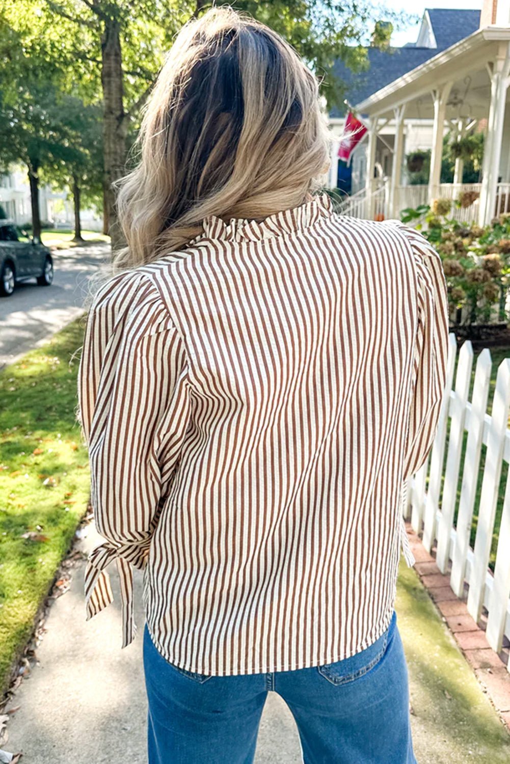Person wearing a striped shirt and blue jeans walking on a sidewalk with a house and flag in the background.