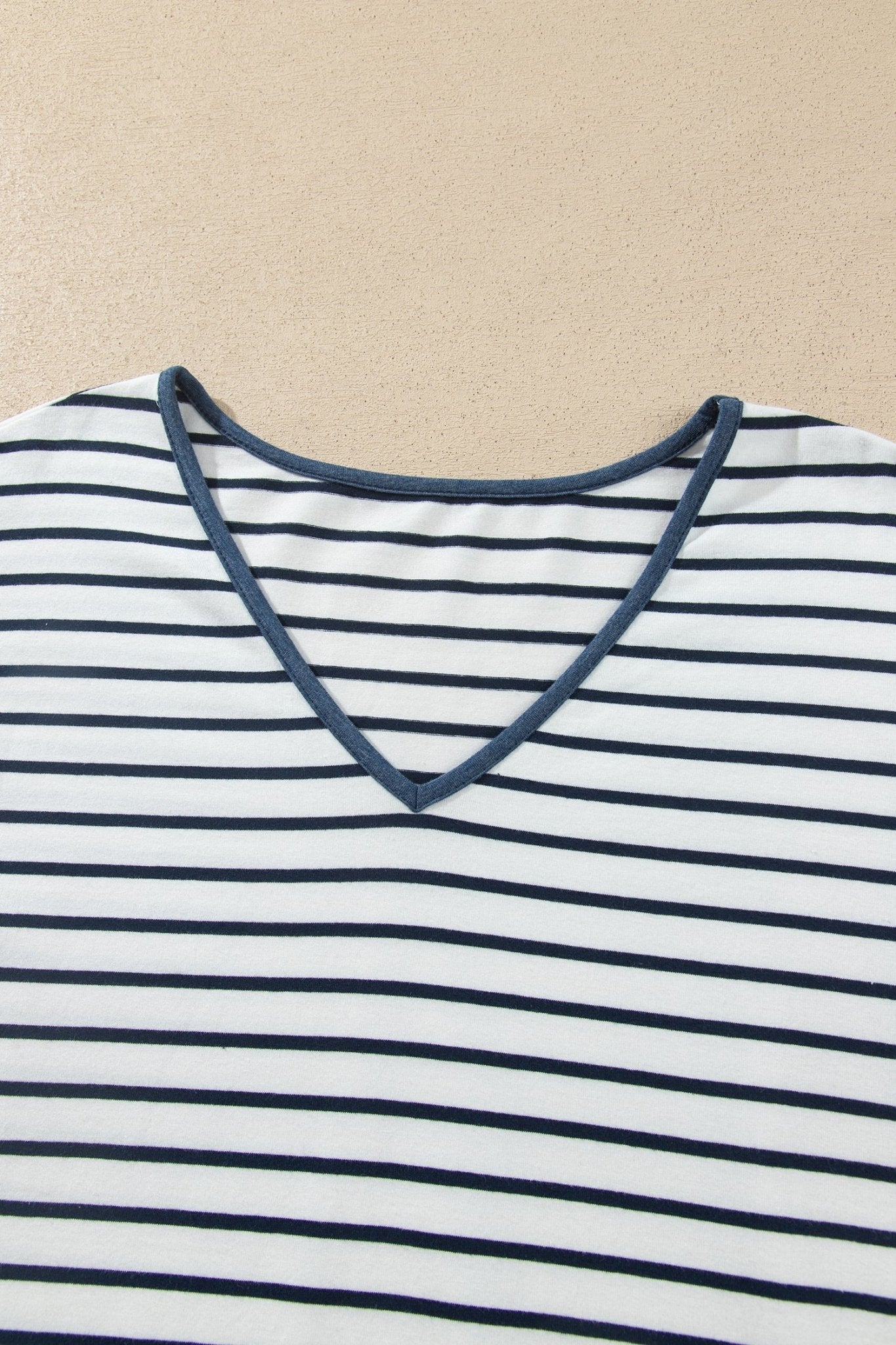Close-up of a white V-neck top with thin navy stripes and solid navy trim on beige.