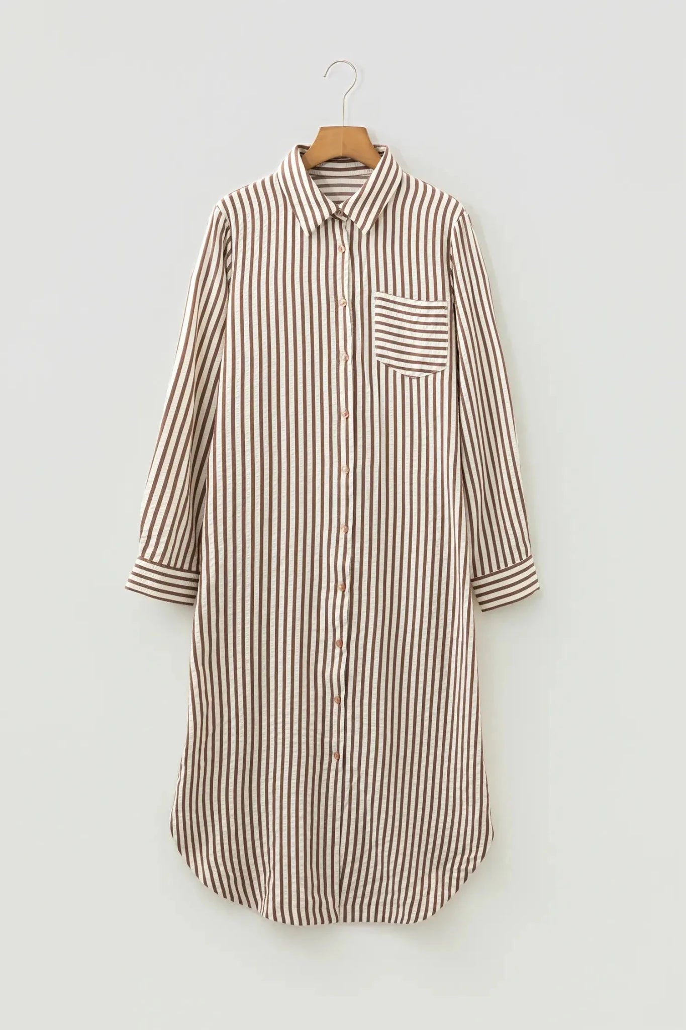 A long-sleeve button-front shirt dress with vertical brown and beige stripes, featuring a single chest pocket, shown hanging on a wooden hanger against a light gray background.
