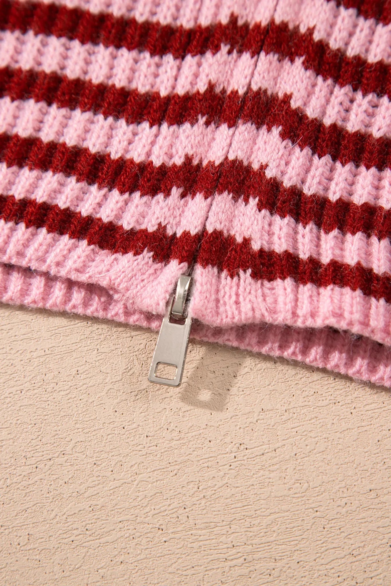 Close-up of pink-dark red striped knit garment with silver zipper on beige surface.