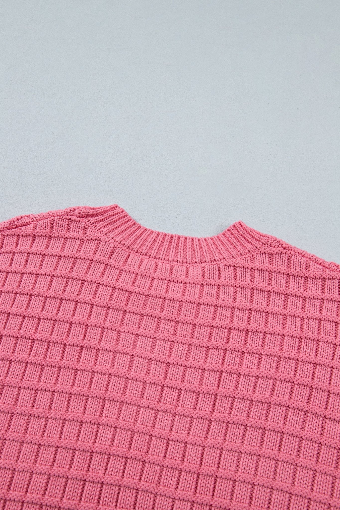 Close-up of a pink ribbed knit sweater showing the crew neck and textured waffle pattern against a light gray background.