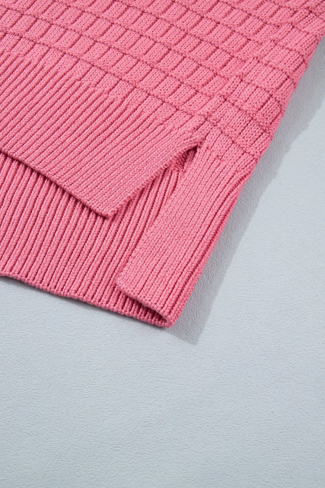 A close-up of a pink ribbed knit garment corner, showing the texture and folded hem detail against a light gray background.