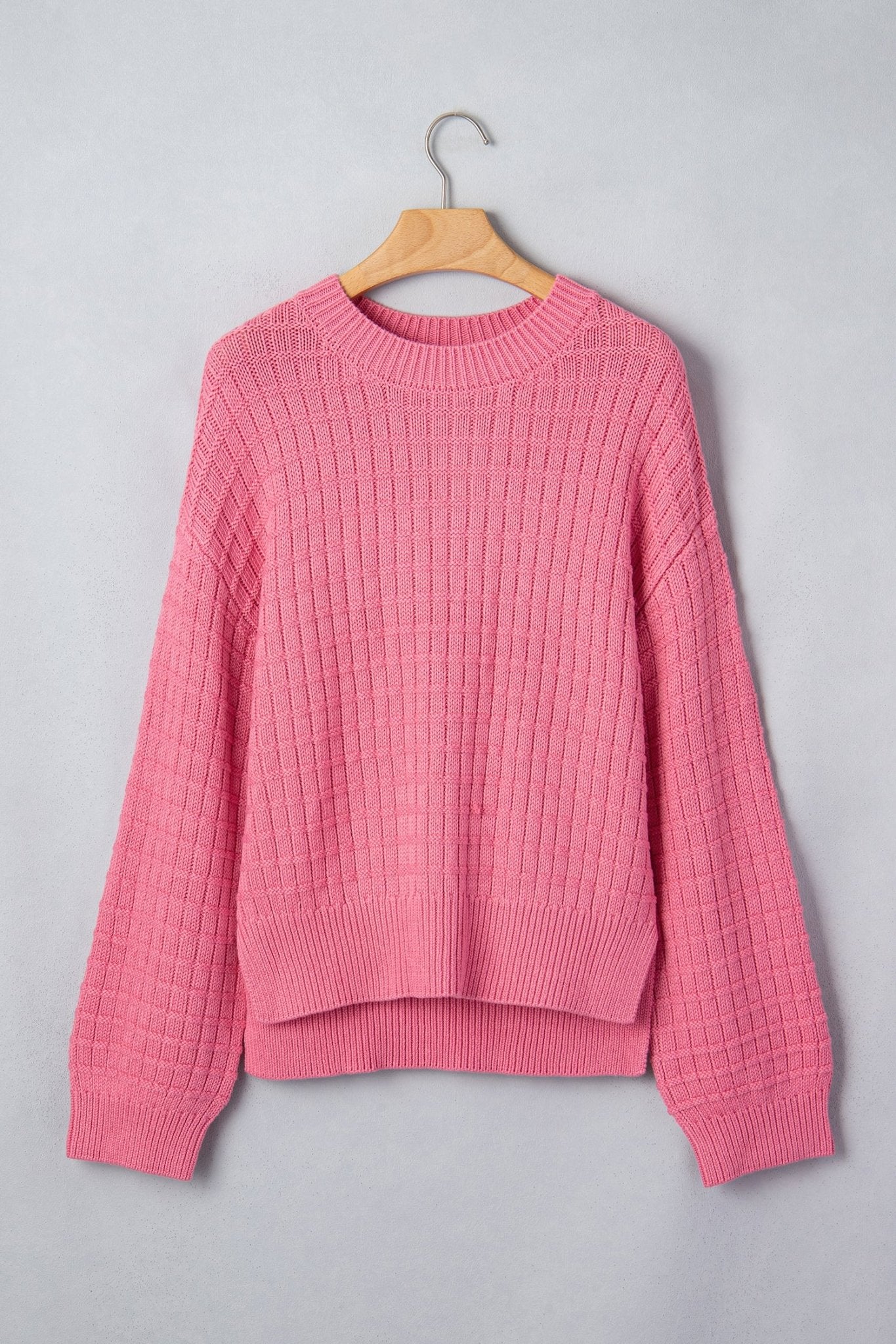 A pink textured knit sweater with a crew neck and ribbed cuffs and hem, hanging on a wooden hanger.
