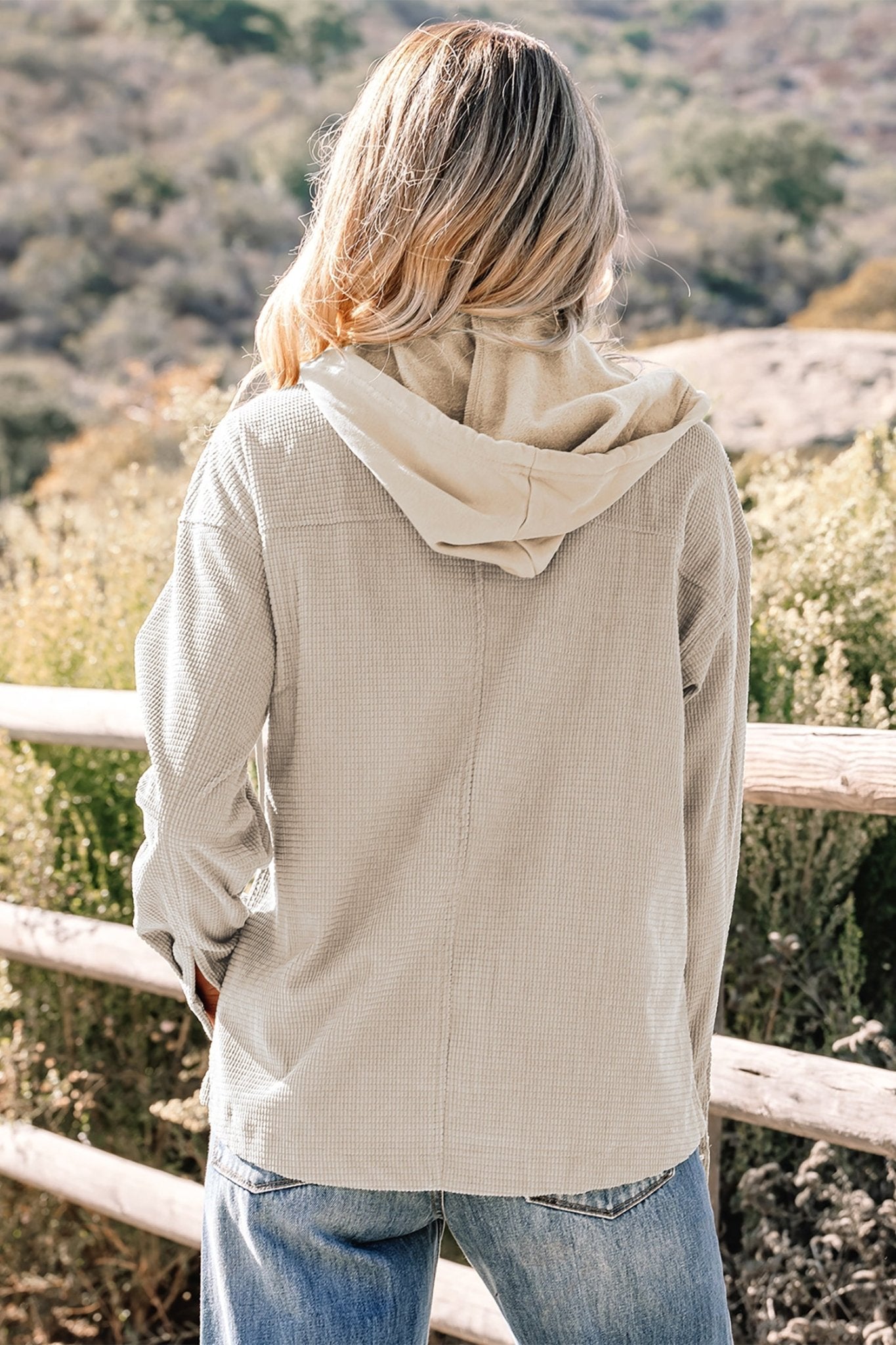 Person wearing a beige hoodie and jeans standing in a natural setting with trees and a wooden fence.