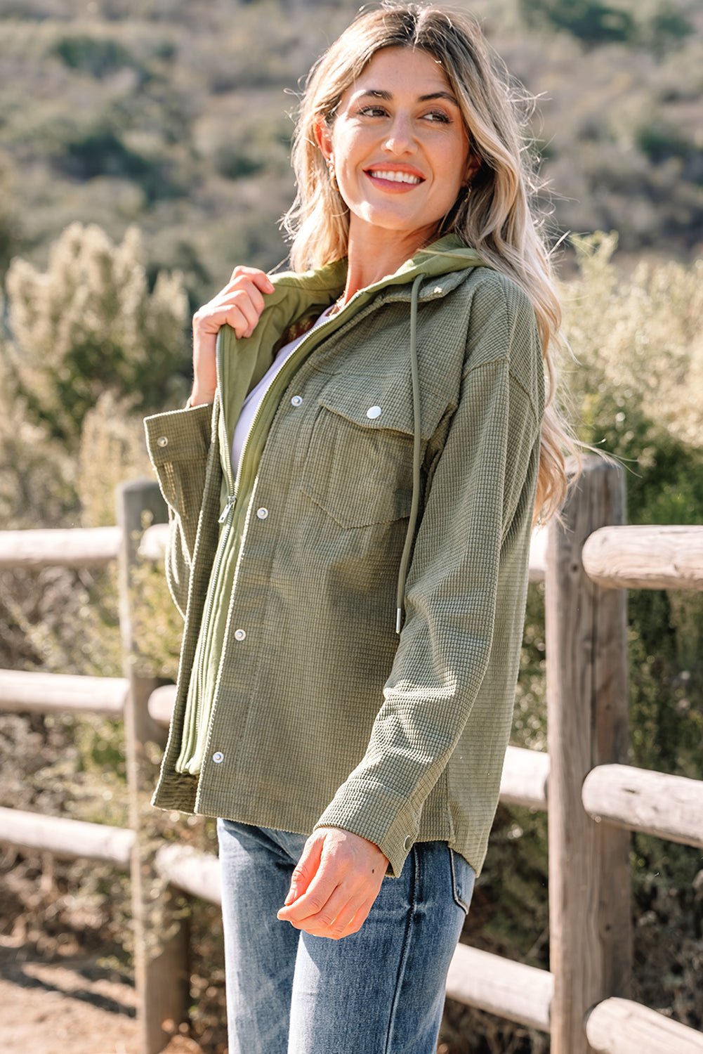 Woman in an olive green jacket outdoors against a wooden fence and natural background.
