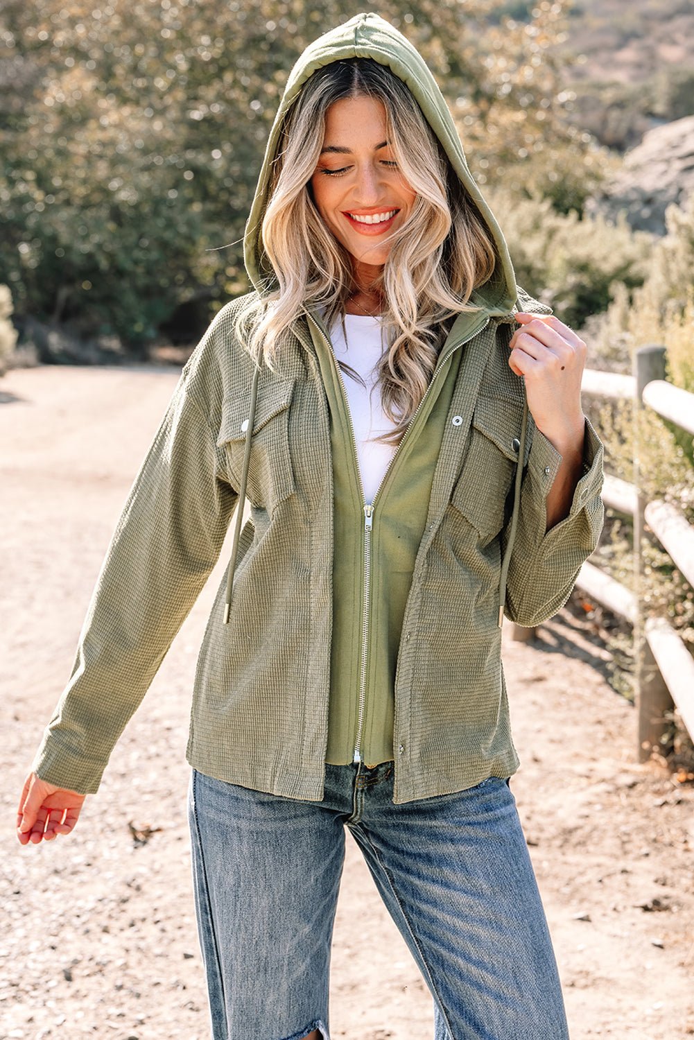 Woman wearing a green hooded jacket outdoors with trees and a fence in the background