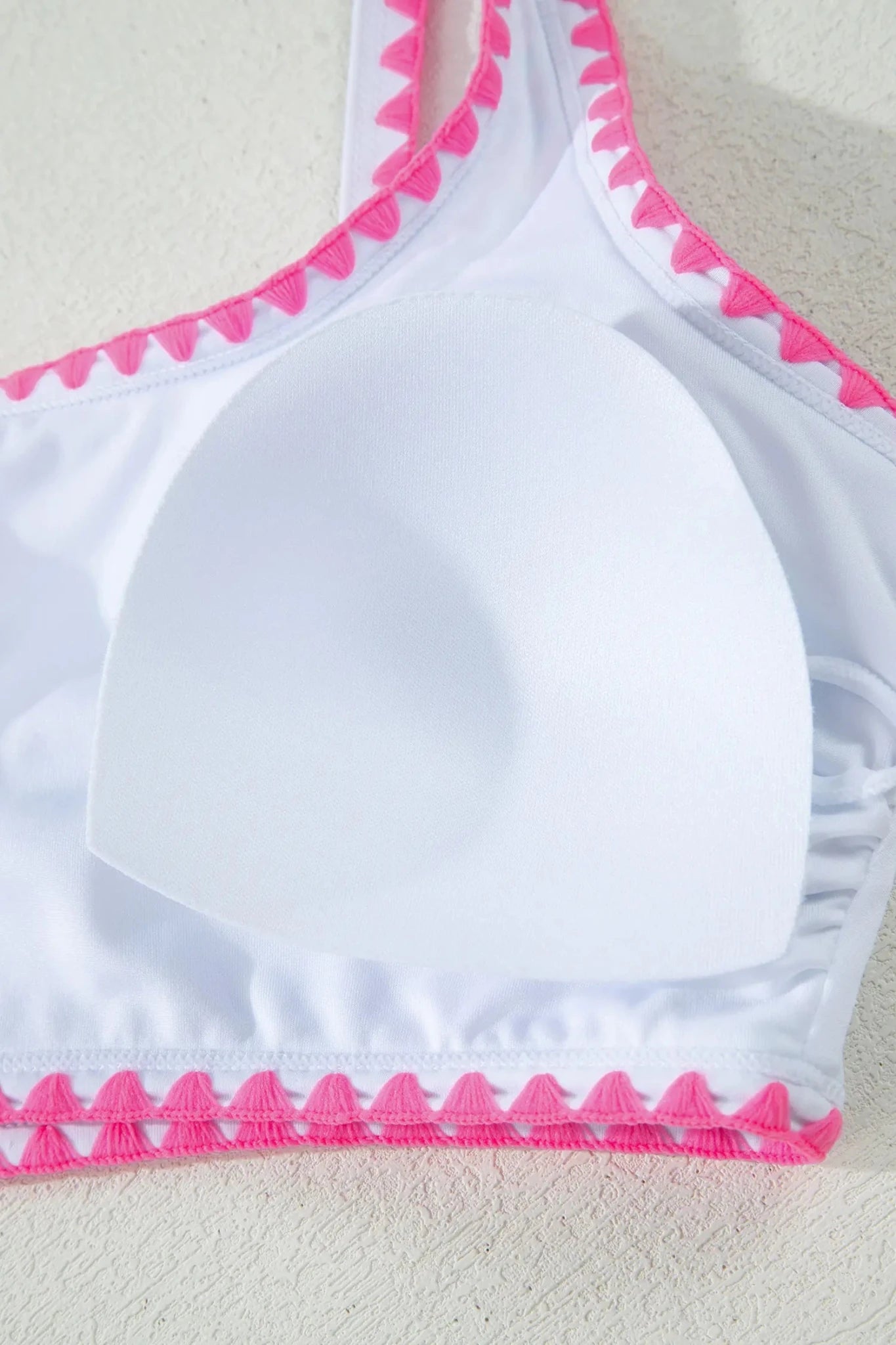 White bikini top with bright pink scalloped trim along the neckline and hem, shown close up on a light textured background.
