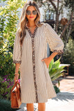 A woman wearing a beige and white vertically striped midi dress with leopard print trim on the collar, placket, and cuffs, holding a brown handbag in an outdoor garden setting.