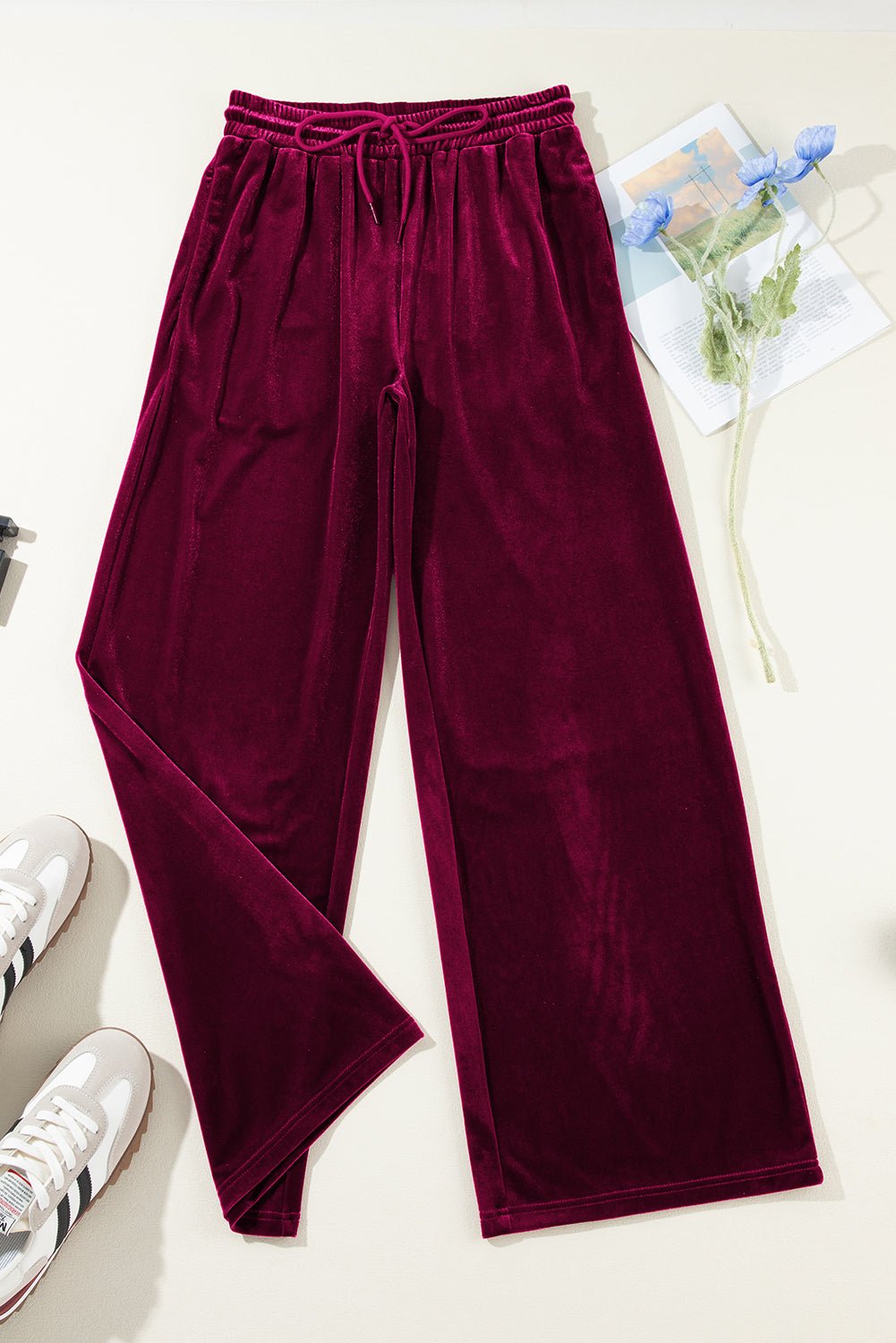 Maroon velvet pants on a light surface with sneakers and a plant in the background