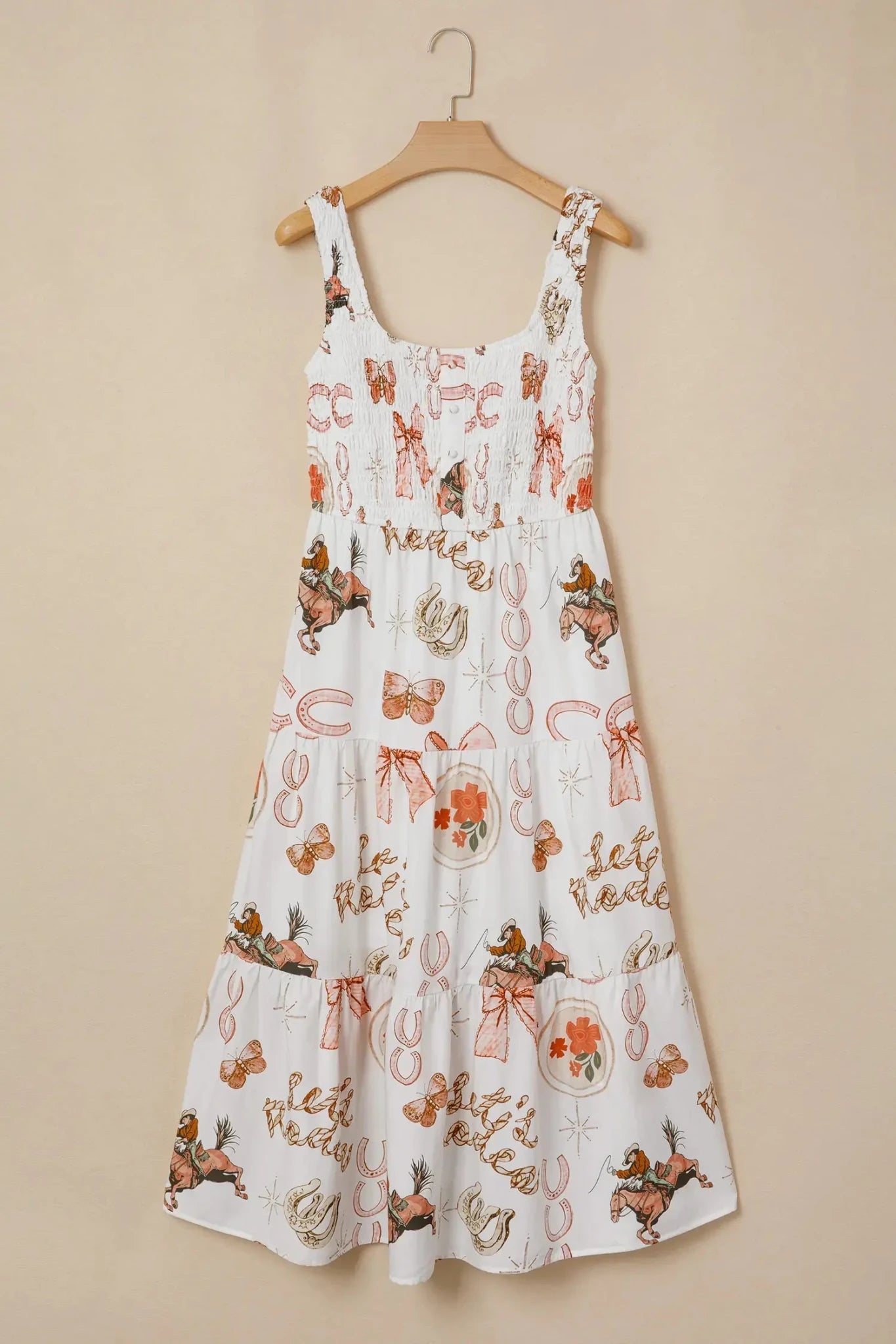 A white sleeveless sundress with a smocked bodice and tiered skirt, featuring an all-over whimsical print of fairies, bows, and autumn motifs in orange, brown, and beige, hanging on a wooden hanger against a solid beige wall.