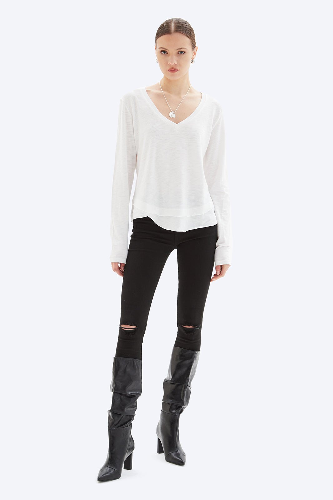 Woman wearing a white long-sleeve top, black ripped jeans, and black knee-high boots on a white background