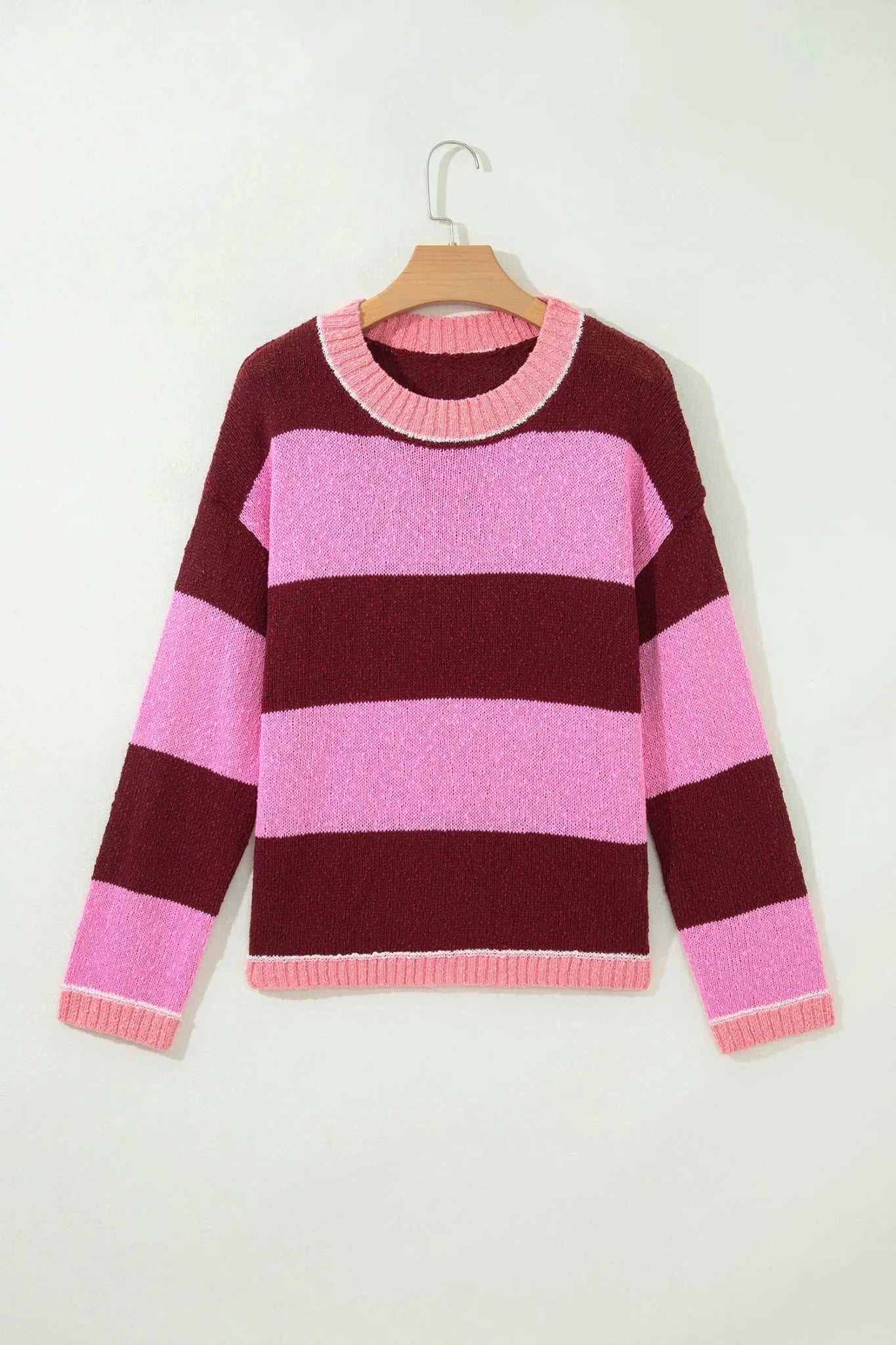 A pink and burgundy striped knit sweater hanging on a wooden hanger against a plain off-white wall.