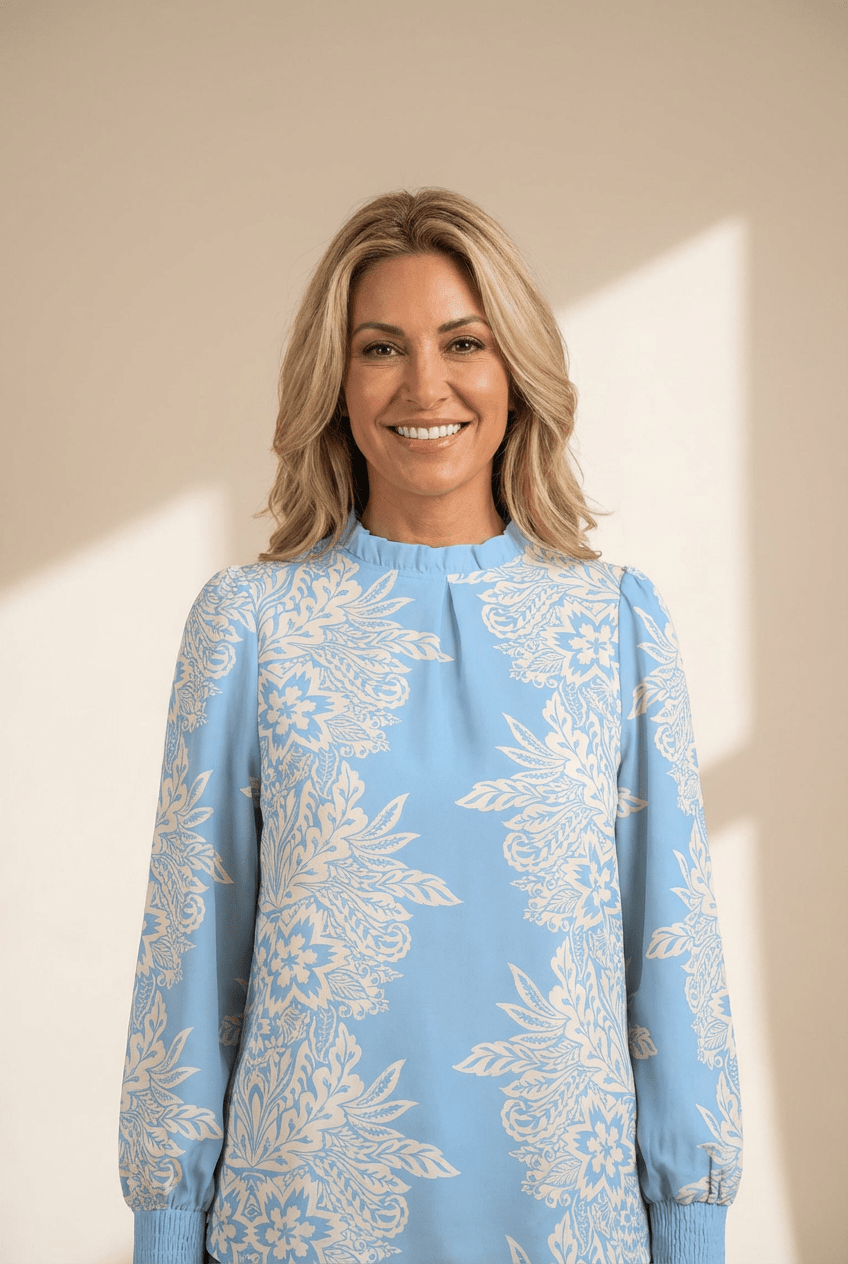 Smiling blonde woman wearing light blue floral blouse against a warm beige background with soft sunlight.