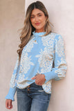 Woman wearing light blue floral knit top with ruffled mock neck and blue jeans against a beige wall.