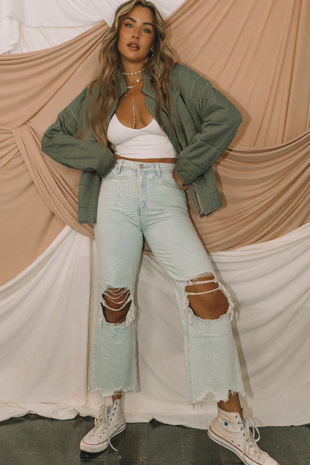Woman in a green jacket, white top, and ripped light blue jeans against draped curtains.
